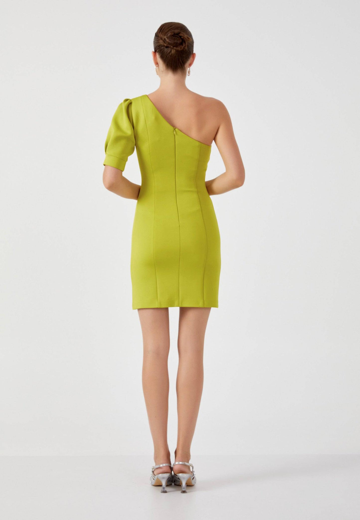 Back view of a woman in a lime green asymmetrical dress, showcasing the fitted silhouette and back zipper closure, standing gracefully with her hair in a sleek bun.