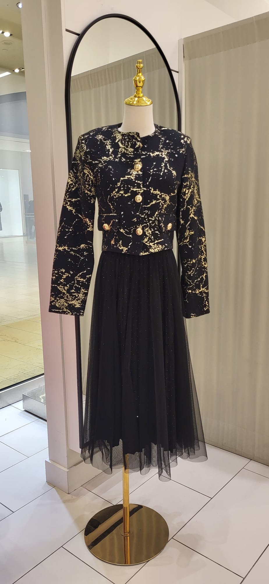 A chic black and gold tweed blazer with gold buttons, styled over a black tulle skirt, displayed on a mannequin in a fashion boutique.